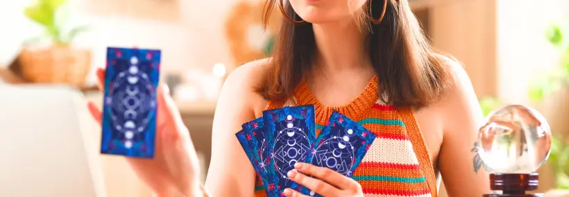 Woman wearing a colorful striped top holding blue tarot cards in both hands, with a crystal ball visible on the table beside her.