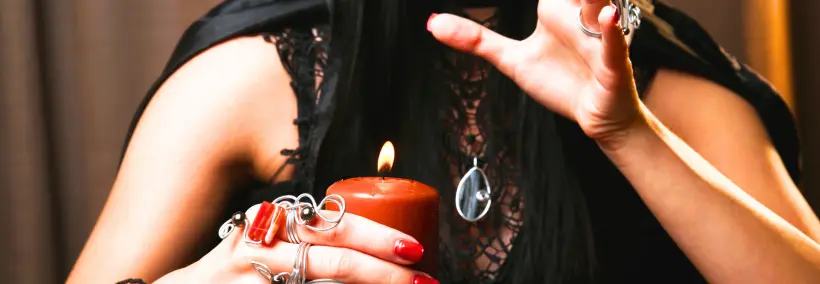 Hands holding a dark crystal or stone object above a lit red candle, creating an atmospheric mystical scene.