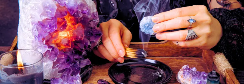 Hands performing a ritual or divination practice over a dark scrying bowl, surrounded by purple amethyst crystals, lit candles, and mystical items with smoke or incense visible.