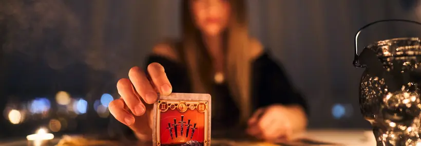 A person's hand holding a small red tarot or oracle card with decorative symbols, set against a softly lit background with candles and bokeh lights creating an atmospheric