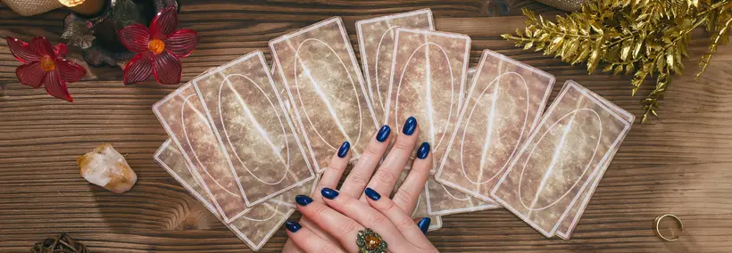Hands with dark blue nail polish positioned over a spread of tarot cards arranged in a fan pattern on a wooden surface, surrounded by decorative elements including red flowers, crystals, and golden tinsel.
